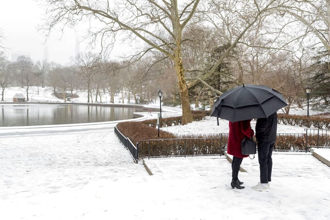 A couple huddle under an umbrella with the snow-covered Park surrounding Conservatory Water behind them