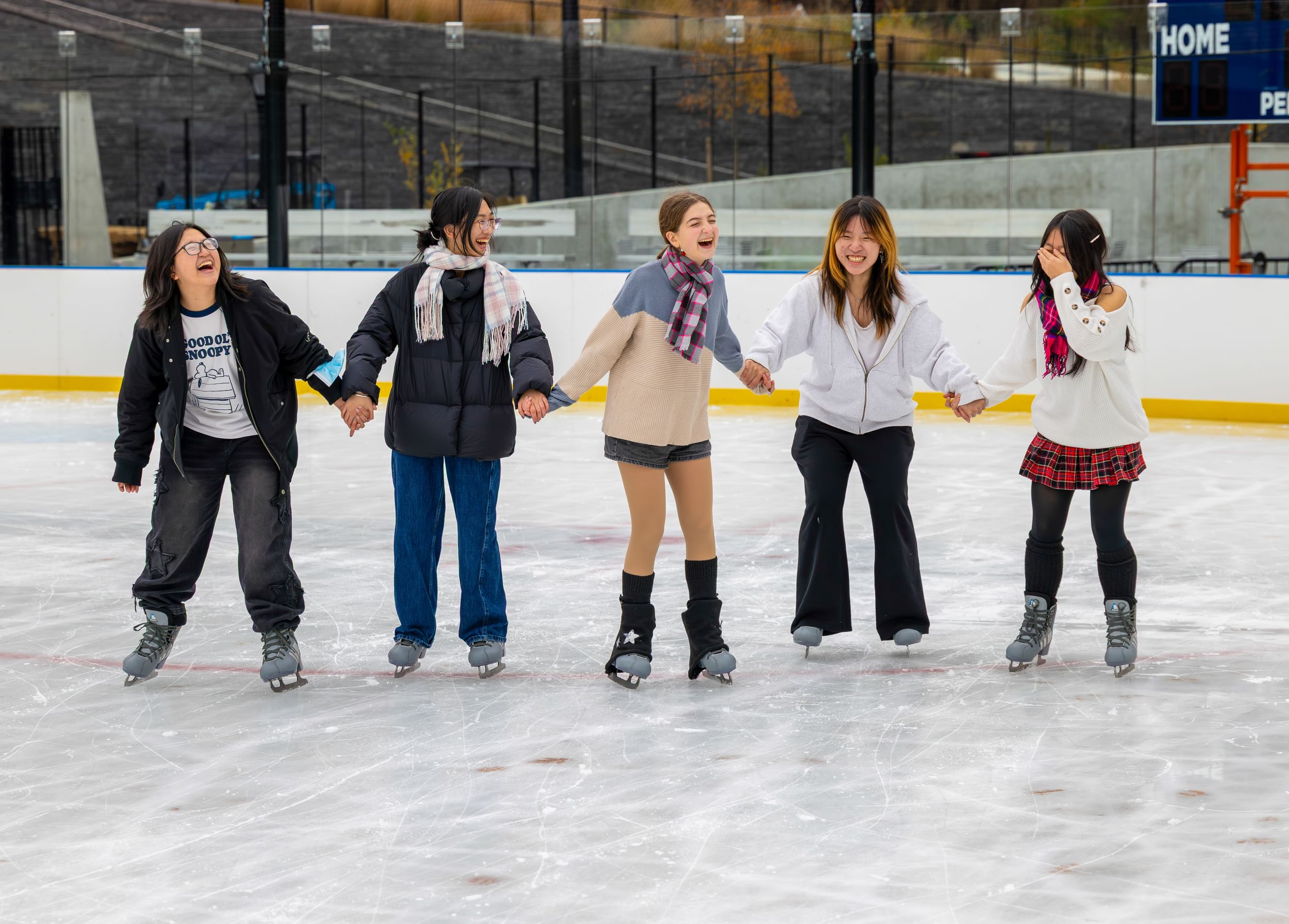 A group of young people hold hands, laughing, while ice-skating together.