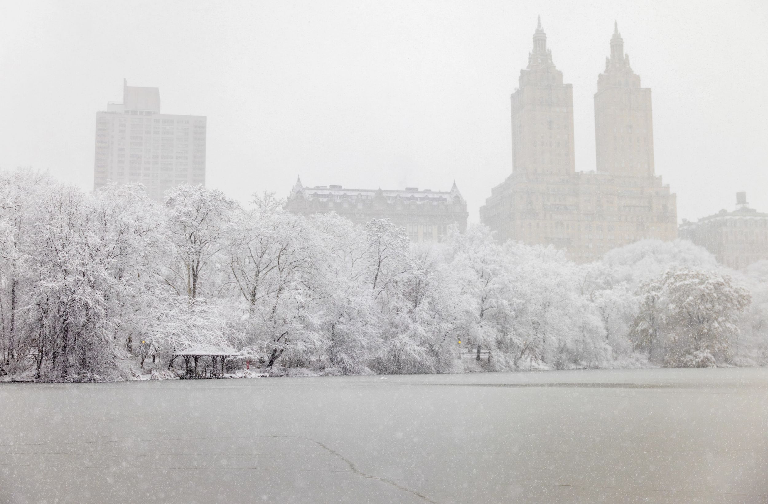 Lake in Central Park in Winter, ice covering the water and snow on trees and tall buildings in the background