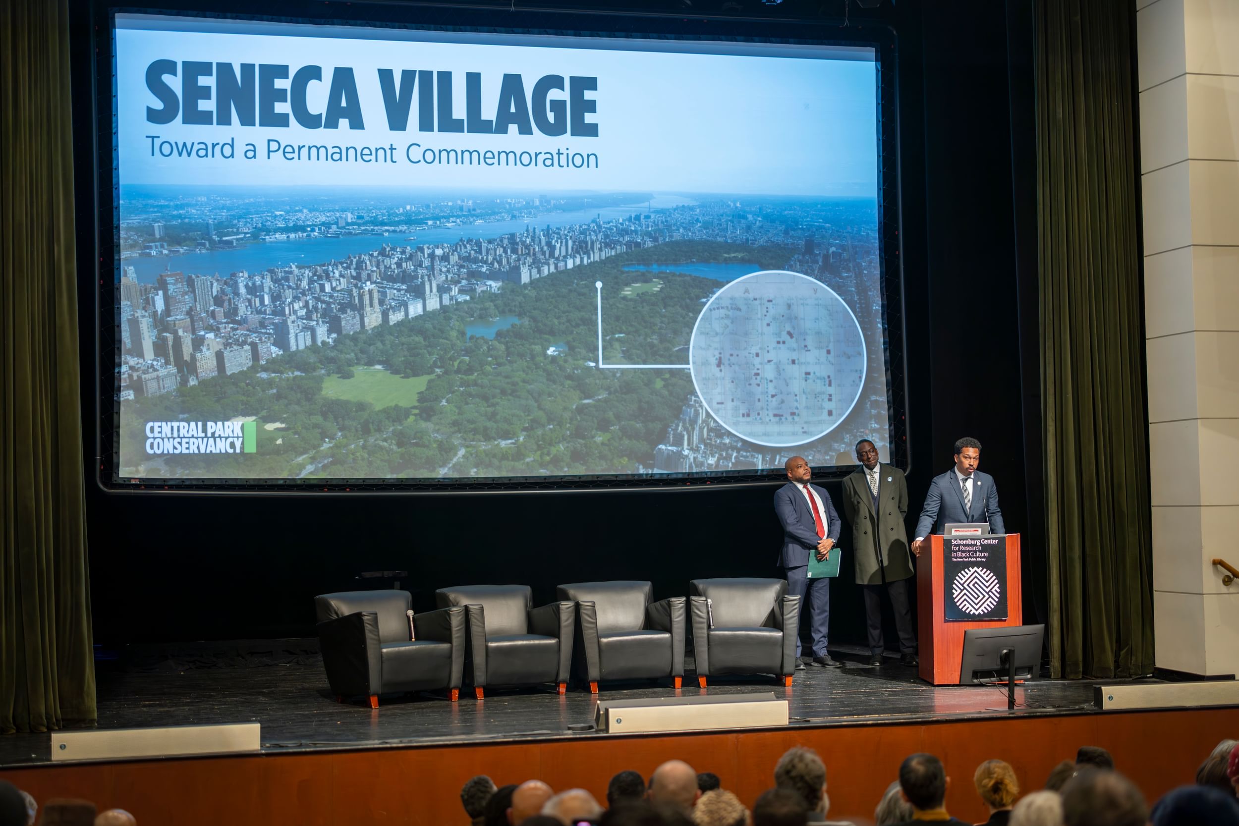 Three Black men stand at the podium at the Schomburg Center for Research in Black Culture in Harlem. On the screen behind them is a photo of Central Park with a historical map of Seneca Village.