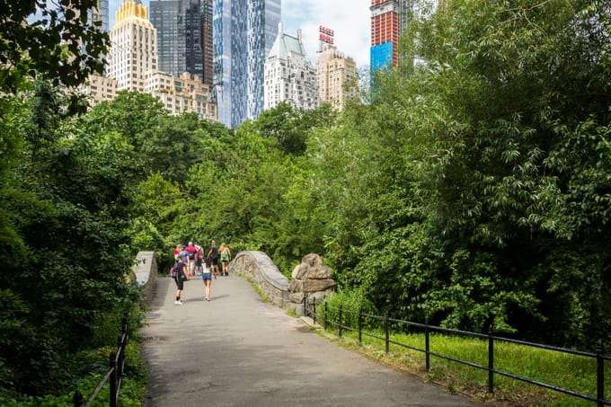 Park goes walk across a bridge that spans the Pond, with Fifth Avenue's buildings looming in the background